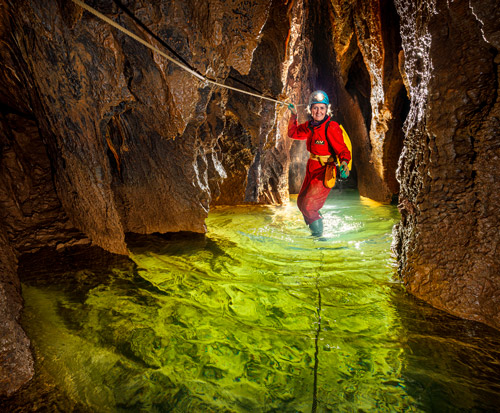 Image of female caver wearing red-overalls holding and standing on wires through brightly lit water in a cave